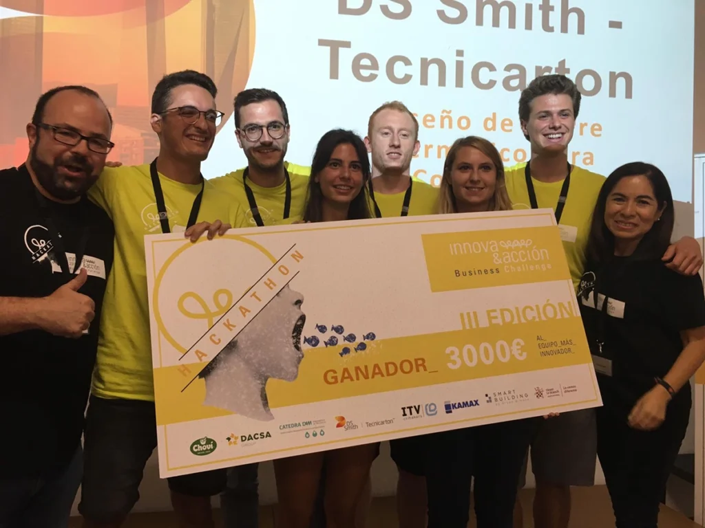 Equipo de participantes posa junto a Juan Pastor Bustamante con un cheque de premio en un escenario tras un hackathon.