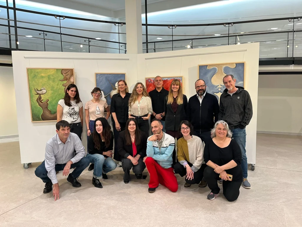Foto de grupo de varias personas posando, entre ellos Juan Pastor Bustamante, en una sala tipo galería con paneles y cuadros al fondo.