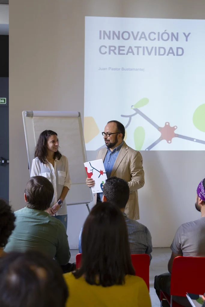 Sesión de formación en creatividad e innovación con Juan Pastor Bustamante como ponente y participantes, con proyector y rotafolios al fondo.