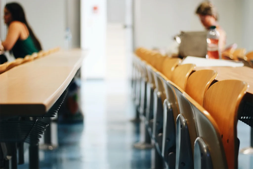 Aula con mesas y filas de sillas, con estudiantes desenfocados al fondo, relacionada con orientación académica y profesional.