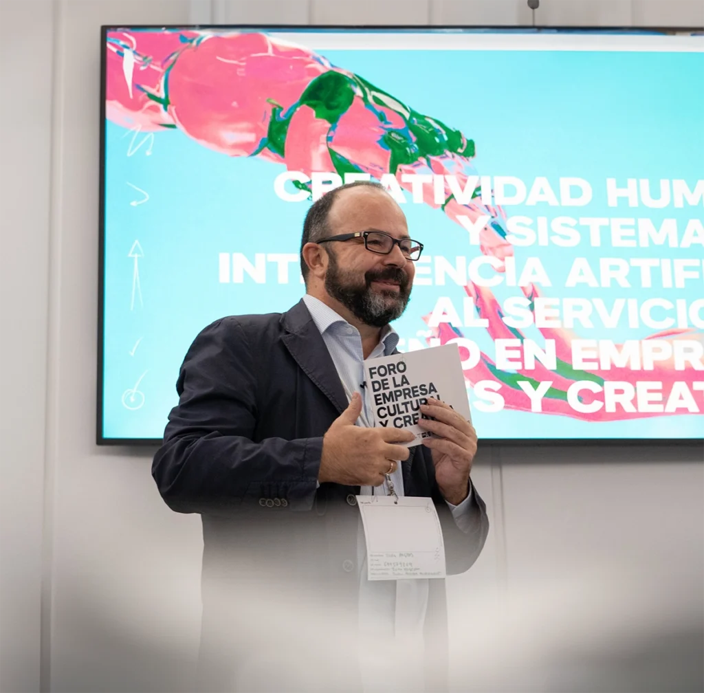 Juan Pastor Bustamante sosteniendo un folleto, con pantalla de creatividad e IA al fondo.