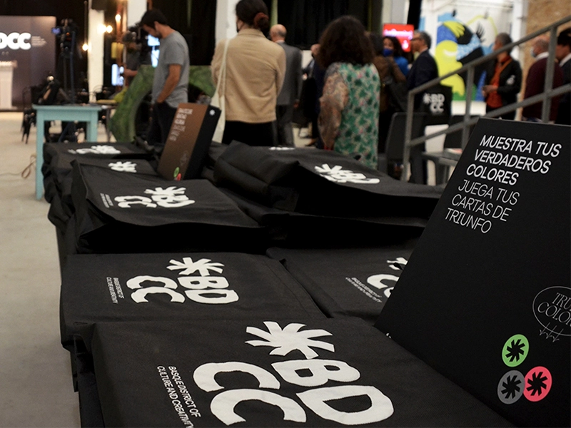 Mesa con bolsas BDCC y cartel informativo en un evento, para programa de consolidación empresarial.