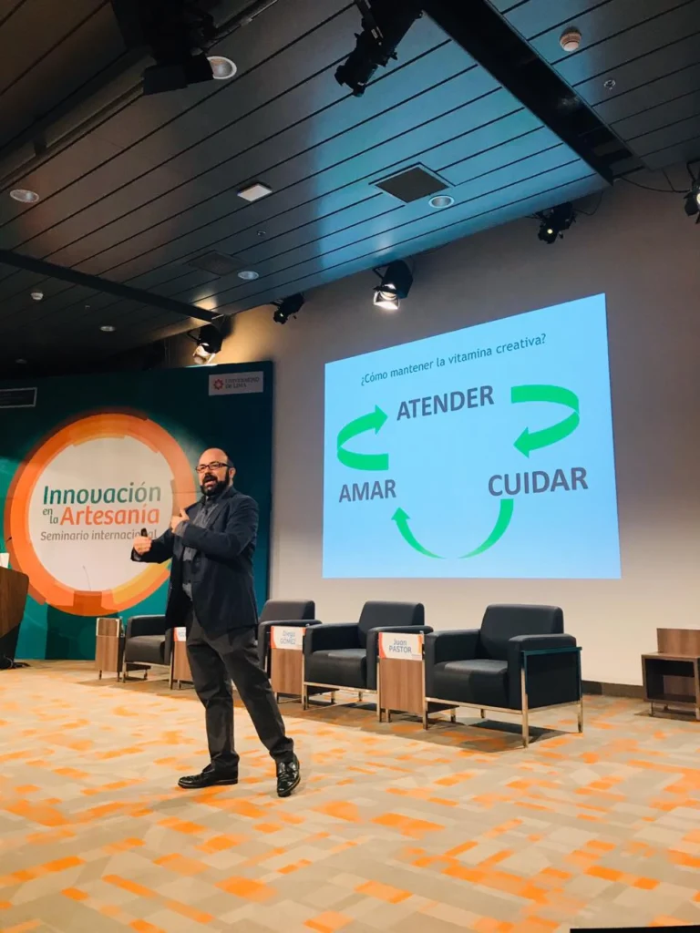 Juan Pastor Bustamante en escenario durante una conferencia, con diapositiva proyectada sobre creatividad e innovación.