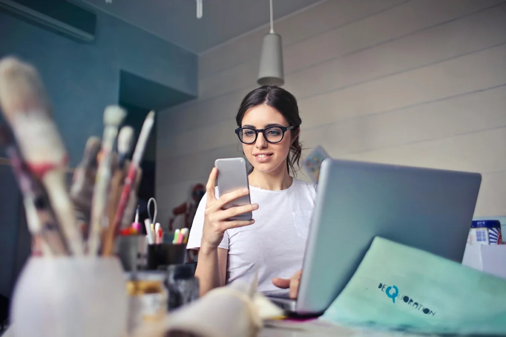 Emprendedora creativa con gafas usando móvil y portátil en un escritorio con pinceles.